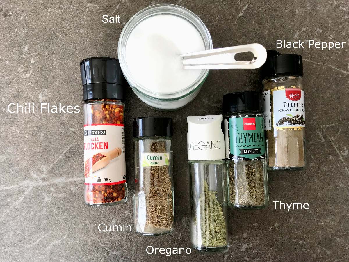 Spices needed for Recipe for Chili con Carne laid out in glass bottles on the table - Black pepper, Thyme, Oregano, Cumin, Chili Flakes, Salt.