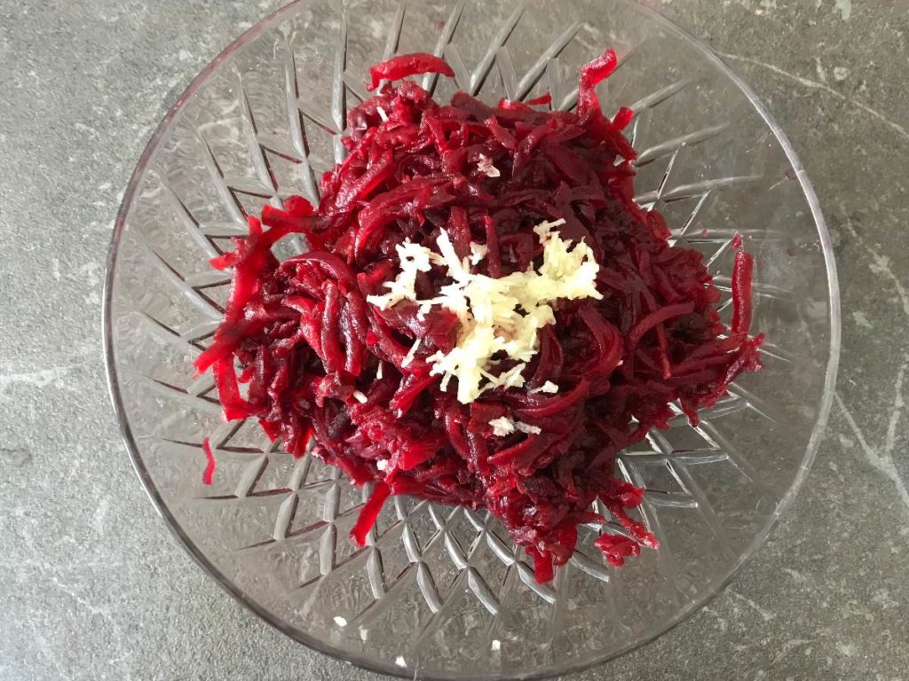 Garlic grated on the top of the grated beets in a glass bowl.