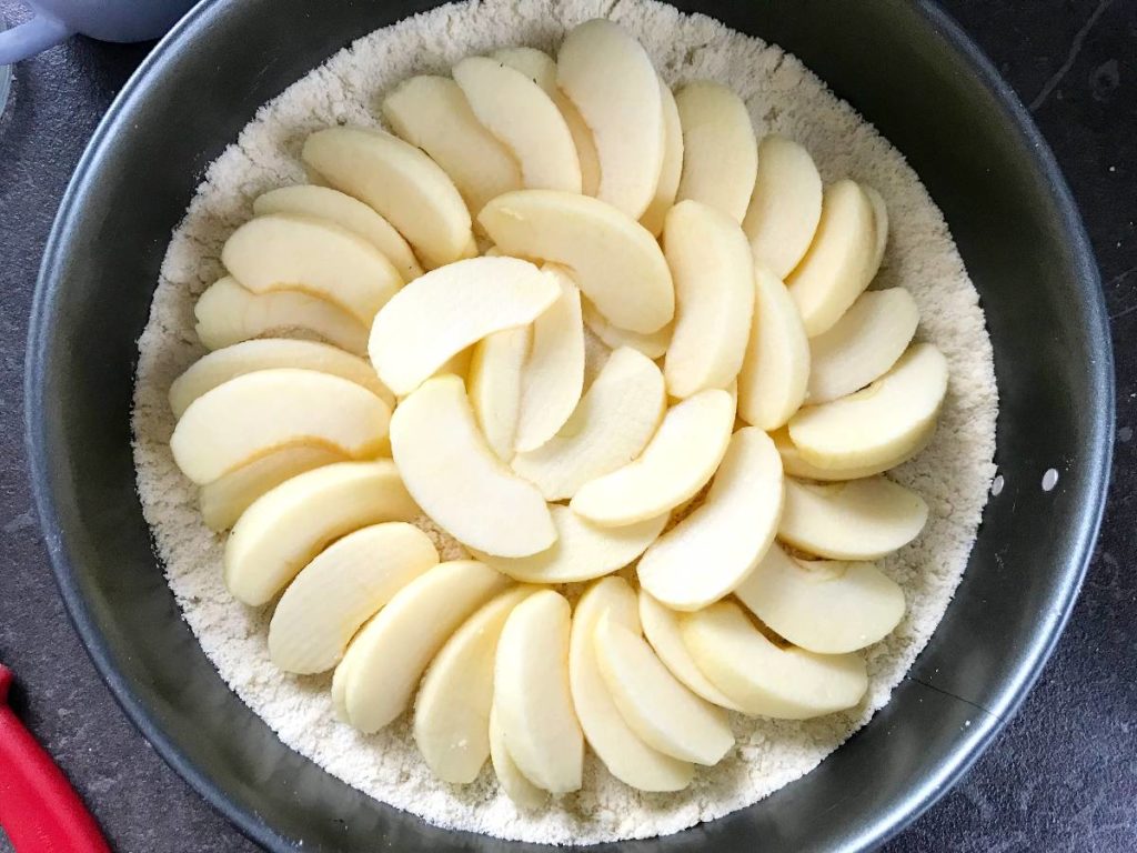 Sliced apples arranged in spring form pan on top of the crumble for cake.