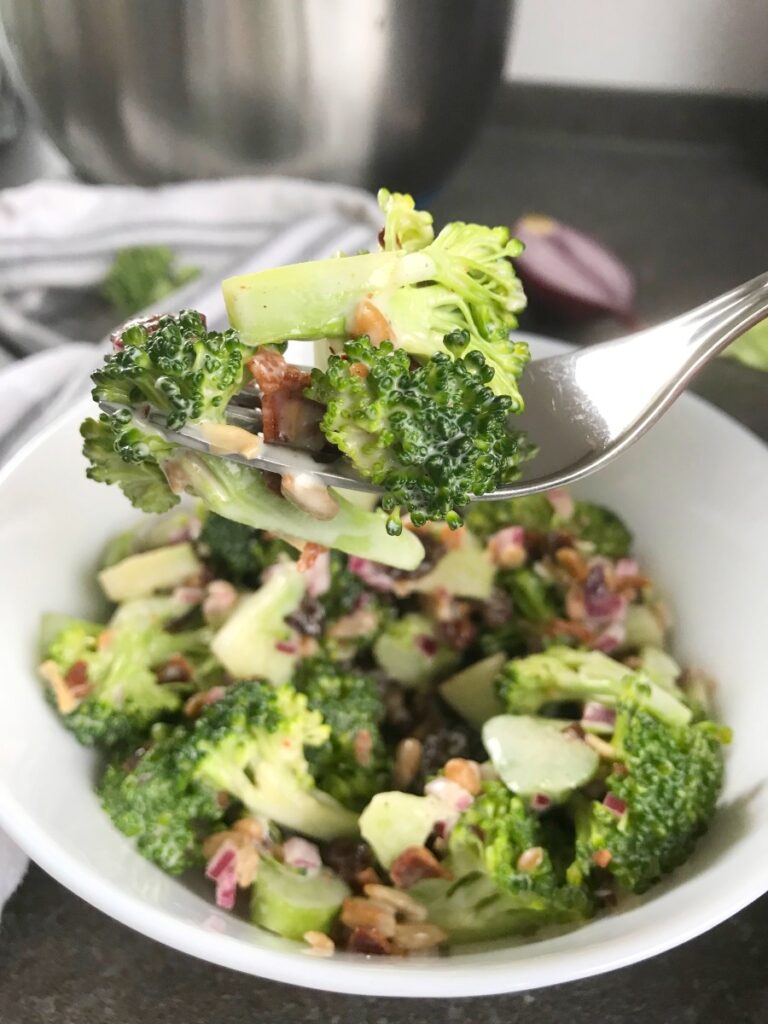 Forkful of Broccoli Cranberry Bacon Salad with the bowl below it and red onion and broccoli pieces all around.