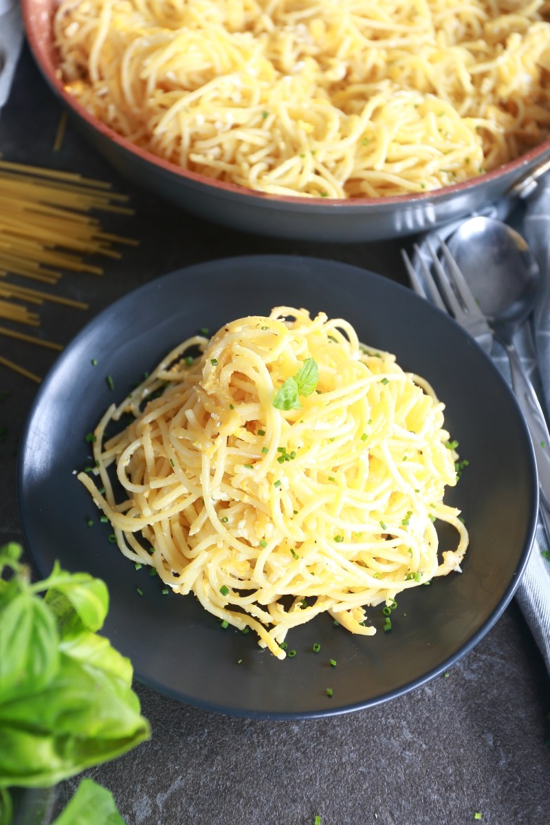 Fried spaghetti on a black plate surrounded by uncooked spaghetti, kitchen towel and basil.