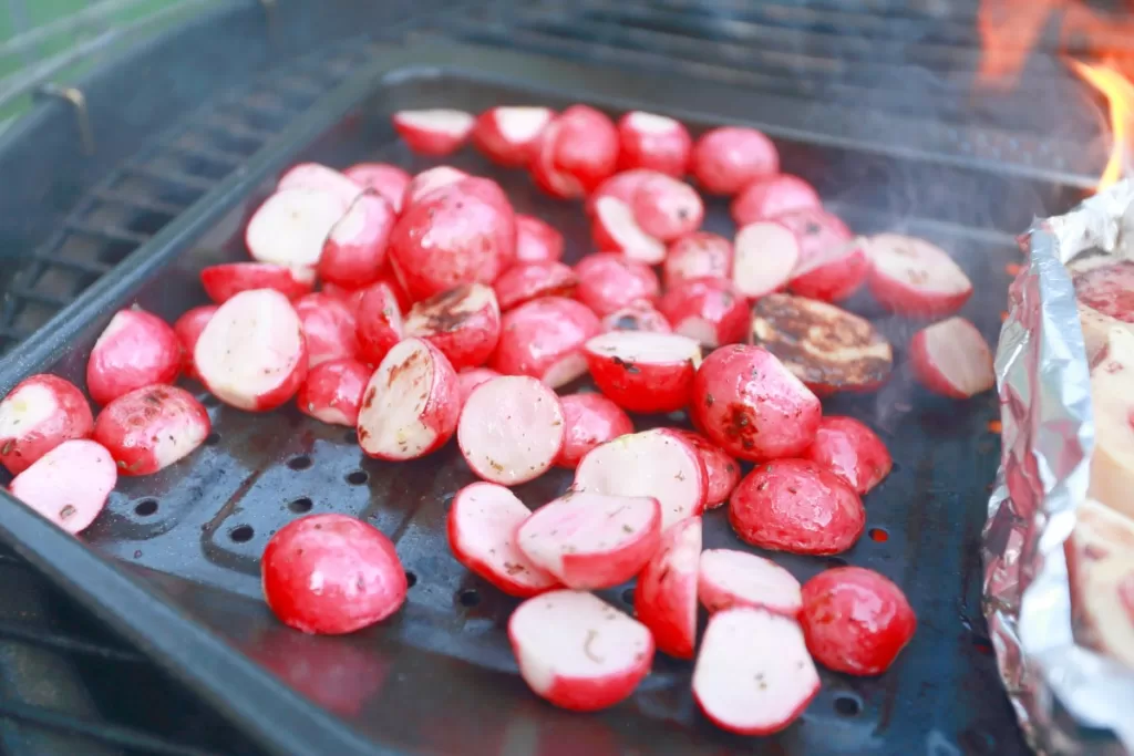 Radishes grilling on a coal grill on a grilling sheet.