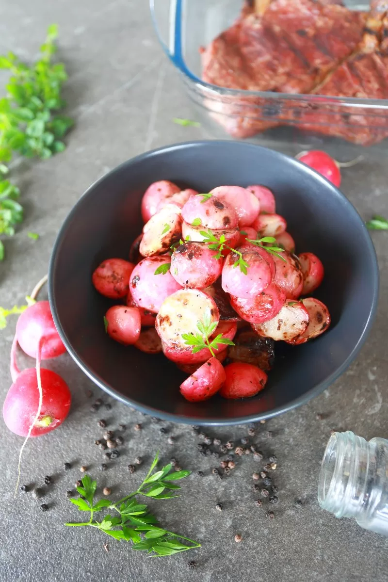 Grilled radishes in a bowl surrounded by fresh radishes and black pepper corns.