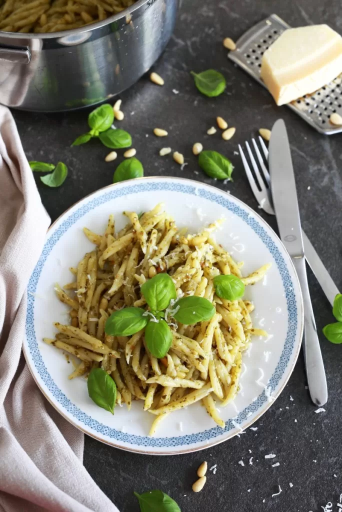 Trofie Pasta served on a plate surrounded by cutlery and sprinkled with basil, Parmesan cheese, and pine nuts.
