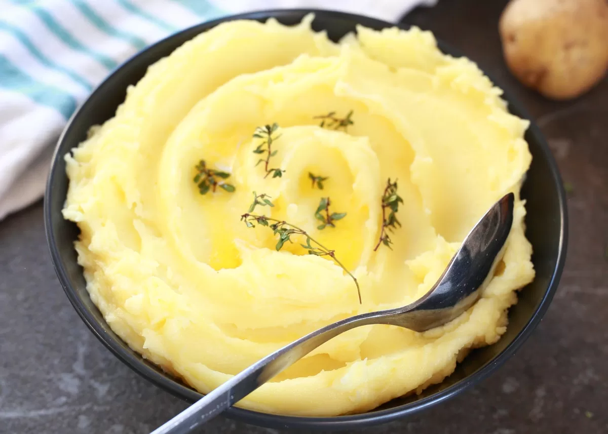 A black bowl filled with creamy mashed potatoes, garnished with a few sprigs of fresh thyme and a drizzle of melted butter. A spoon rests in the mashed potatoes, and a striped cloth and whole potatoes are visible in the background.