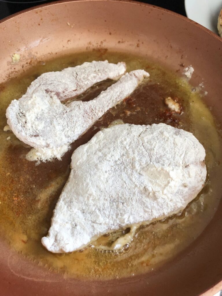 Chicken breast dredged in flour frying in a skillet.