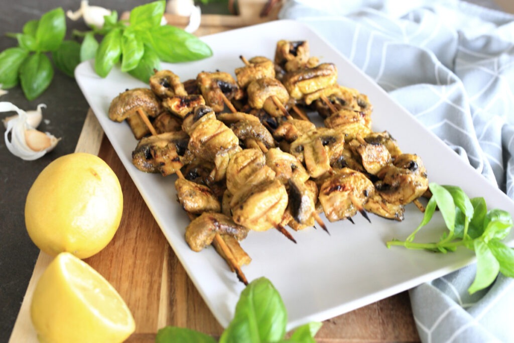 Grilled mushrooms on skewers on a platter surrounded by greens and lemon.